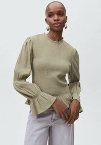 Olivno zelena, teksturirana majica s napuhanim rukavima i nabranim ovratnikom; uparena s svijetlosivim trapericama. Ima flared manžetne i prilagođen struk.