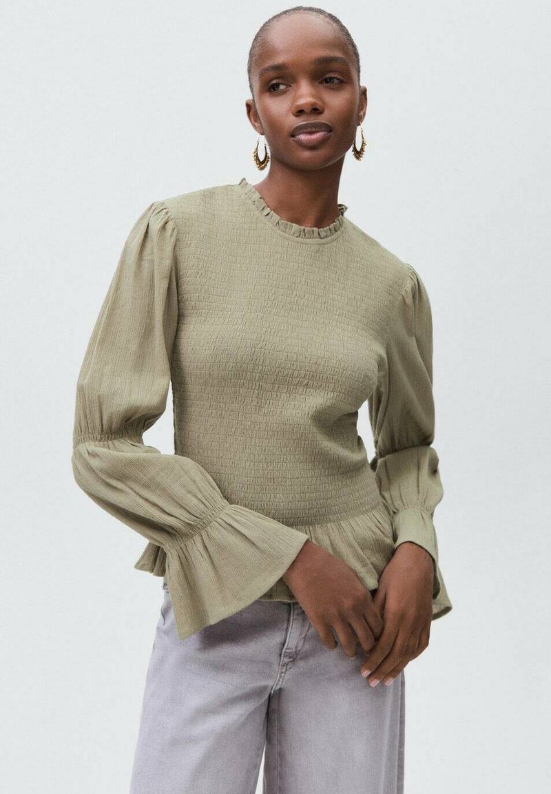 Olivno zelena, teksturirana majica s napuhanim rukavima i nabranim ovratnikom; uparena s svijetlosivim trapericama. Ima flared manžetne i prilagođen struk.