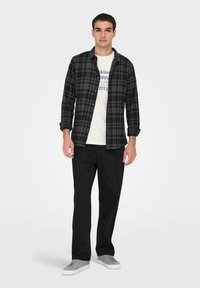 Heren zwart en grijs geruite flanellen overhemd over een witte graphic t-shirt, gecombineerd met zwarte broek en grijze sneakers. Schoon, casual uiterlijk.