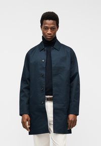 Άνδρας που φοράει σκούρο ναυτικό παλτό με κουμπιά πάνω από μαύρο πουλόβερ με ψηλό λαιμό και ανοιχτόχρωμο παντελόνι με μαύρη ζώνη, στέκεται μπροστά σε λευκό φόντο.