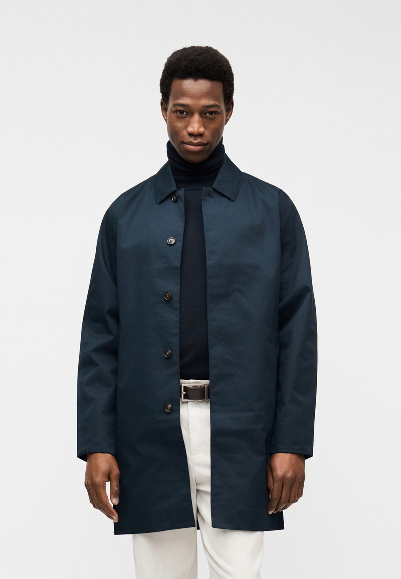 Άνδρας που φοράει σκούρο ναυτικό παλτό με κουμπιά πάνω από μαύρο πουλόβερ με ψηλό λαιμό και ανοιχτόχρωμο παντελόνι με μαύρη ζώνη, στέκεται μπροστά σε λευκό φόντο.