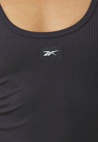 Top de tirantes negro con canalé que presenta un escote redondo y un parche con el logo en la parte frontal. La tela parece elástica y estructurada.