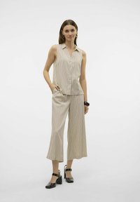 Camisa sin mangas beige con rayas verticales, con cuello y botones al frente. Combinada con pantalones de pierna ancha a rayas y zapatos negros de tacón.