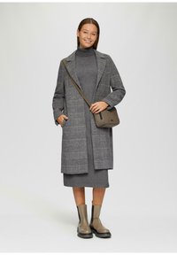 Lange grijze jas met een ruitpatroon, grijze coltrui jurk en bruine enkellaarsjes. Een bruine schoudertas completeert de outfit.
