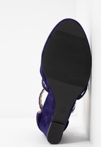 Chaussure à talon haut en daim violet avec une semelle en caoutchouc noire texturée, présentant un design découpé et une fine bride autour de la cheville.