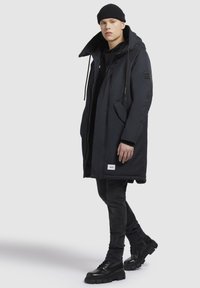 Joven con un gorro negro, abrigo largo acolchado negro con capucha, jeans negros y botas de cuero gruesas negras, de pie sobre un fondo blanco.