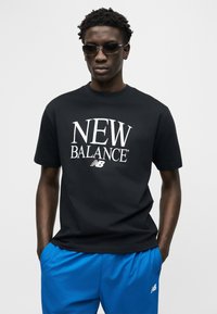 Άνδρας με μαύρο T-shirt New Balance και μπλε σορτς, με τα χέρια στις τσέπες, ποζάροντας μπροστά σε απλό φόντο.