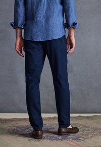 Pantaloni din in bleu marin cu croi drept, asortați cu o cămașă din chambray albastru deschis, văzuți din spate, purtați cu pantofi maronii.
