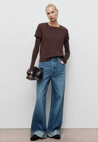 Bruine lange mouwen shirt gecombineerd met een wijd uitlopende blauwe jeans, met een omgeslagen zoom. Het model heeft een kleine zwarte handtas in de handen.