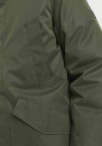 Zelena izolirana jakna s gladko teksturo, žepom na prsih in poševnim stranskim žepom z vstavljenim detajlom.