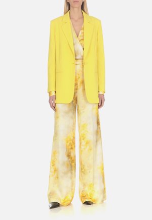 Blazer giallo con revers sopra una tuta a fantasia gialla chiara con gamba ampia. Tessuto morbido con una texture liscia e dettagli lucidi.