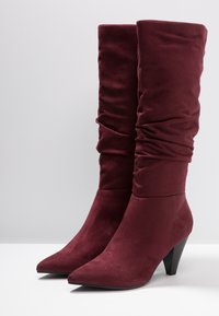 Un par de botas de ante burdeos de media caña con punteras puntiagudas y tacones cónicos apilados sobre una superficie blanca con un fondo liso.