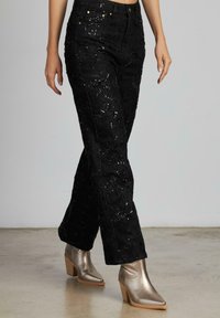 Jeans neri ricamati con paillettes e taglio dritto, abbinati a stivaletti metallizzati con tacco, che mostrano dettagli testurizzati.