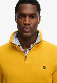 Żółty sweter z dzianiny z zamkiem błyskawicznym, prążkowanym kołnierzem i haftowanym logo na klatce piersiowej. Wewnętrzna część kołnierza ma niebieskie i białe paski.