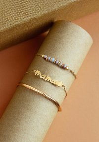 Tres pulseras sobre un tubo de papel: una cadena dorada con "mama", una pulsera de cuentas coloridas y un brillante brazalete dorado. Texturas suaves y materiales variados.