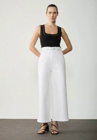 Schwarzes Tanktop mit rundem Halsausschnitt, kombiniert mit hoch taillierten weißen weiten Hosen mit Gürtel. Schwarze Riemchensandalen vervollständigen den Look.