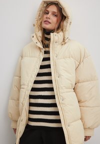 Krémová pufková bunda s kapucňou, so zipsovým predným zapínaním, veľkými prešívanými sekciami a cvokmi. Nosená cez čierno-biely pruhovaný sveter.