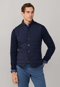 Dunkelblaue gesteppte Weste mit hohem Kragen, Knopfleiste vorne und zwei Vordertaschen, darüber ein strukturiert gestrickter dunkelblauer Pullover mit langen Ärmeln.