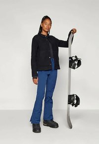 Must must, must särk, sinised suusapüksid ja mustad saapad. Hoides lumelaua ja kinnitustega. Seistes neutraalse tausta ees.