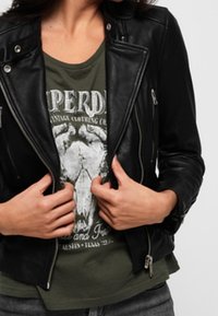 Giacca da motociclista in pelle nera con cerniere argentate e finitura testurizzata, indossata sopra una t-shirt grafica verde con un design di teschio.
