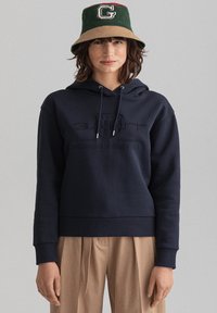 Felpa con cappuccio blu navy con orlo a costine, caratterizzata da un marchio in rilievo. Il modello indossa un cappello a secchiello verde e sabbia, con un logo sulla parte frontale.