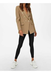 Beżowa marynarka dwurzędowa z złotymi guzikami i dwoma przednimi kieszeniami, w połączeniu z czarnymi legginsami i białymi trampkami. Gładka faktura, dopasowany krój.
