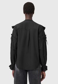 Camicia nera con maniche lunghe, polsini arricciati, dettagli sulle spalle e piega sul retro. Realizzata in un tessuto liscio, progettata per un fit rilassato.