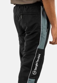 Pantaloni sportivi neri con una vestibilità aderente, dotati di pannelli laterali turchesi, piping bianco e una tasca con zip sul retro. Logo del marchio sulla gamba.