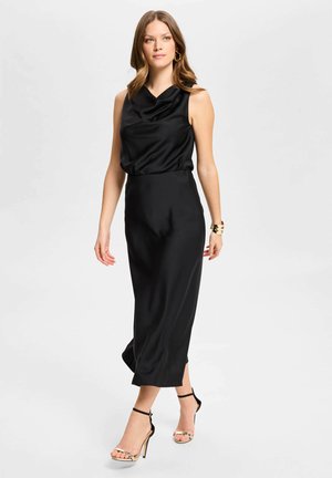 Mujer con vestido negro de satén sin mangas con escote drapeado, pendientes de aro dorados, brazalete de oro y tacones altos negros con tiras.