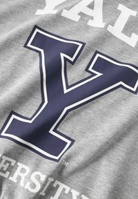 Pelēks sintētiskā materiāla T-krekls ar tumši zilu "Y" logo un baltiem uzrakstiem "YALE UNIVERSITY", kas drukāti uz priekšpuses. Mīksta un gluda auduma struktūra.