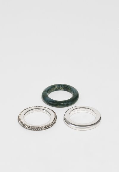 Tres anillos: una banda de resina verde translúcida, una banda de plata con pequeñas piedras claras y una banda de plata pulida con texto grabado.