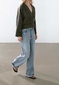 Giacca verde scuro dal taglio ampio con scollo a V profondo, abbinata a jeans larghi a gamba larga azzurri e sandali neri con un leggero accento viola.