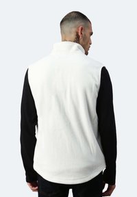 Weißer Fleece-Weste mit hohem Kragen, über einem schwarzen Langarmshirt getragen. Weiche Textur und runder Saum. Die Rückansicht zeigt keine Hardware oder Akzente.
