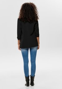 Blazer nero su misura con maniche a tre quarti, abbinato a jeans skinny blu e stivaletti neri alla caviglia. Tessuto liscio con design strutturato.