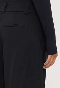 Melnas bikses ar gludu tekstūru, taisnu griezumu, ar aizmugures kabatu un jostas cilpām, apvienotas ar ilgu piedurkņu tumšu blūzi.