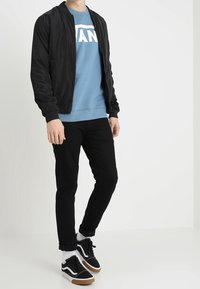 Černá bomber bunda přes světle modrou mikinu s logem "VANS", černé slim fit kalhoty a černé tenisky s bílými detaily a gumovou podrážkou.