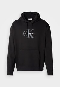 Crna dukserica s kapuljačom od pamuka, s prednjim džepom i otisnutim Calvin Klein logotipom u sivoj i bijeloj boji na prsima.