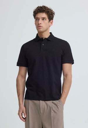 Mężczyzna o kręconych włosach w czarnej, krótkim rękawem koszulce polo i beżowych spodniach z plisami, stojący z jedną ręką w kieszeni.