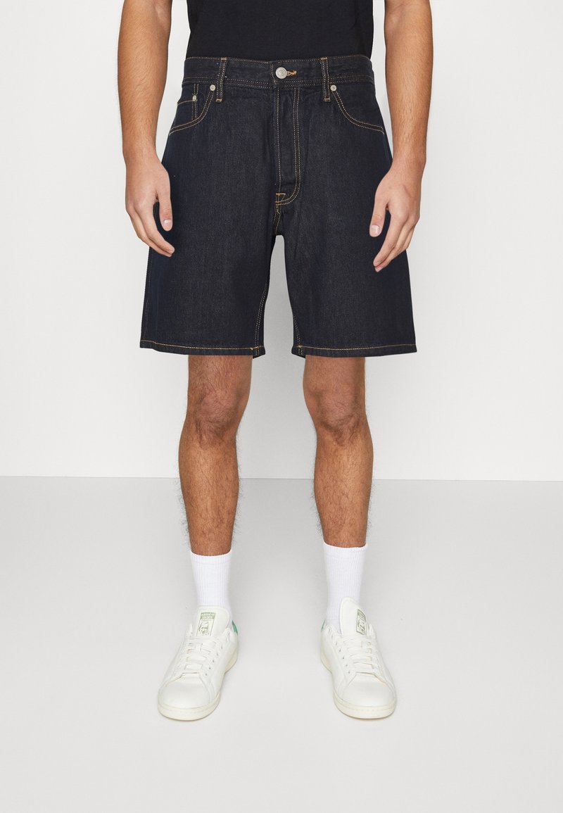 Mörka denimshorts med kontrasterande sömmar, femfickorsmodell och flat front. Bärs med en svart t-shirt och vita sneakers.