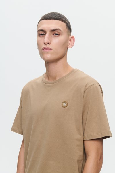 Ung mand med kort, mørkt hår iført en beige T-shirt med et lille rundt logo på brystet, der står foran en ensfarvet lys baggrund.