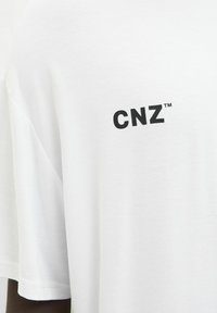 Baltos medvilnės marškinėliai su juodu "CNZ™" logotipu kairėje krūtinės pusėje. Audinys turi lygų, minimalų tekstūrą ir laisvą siluetą.