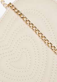 Beżowa torba z teksturowanej skóry z przeszyciami w kształcie serca, wyposażona w pasek z złotym łańcuszkiem. Wzór jest gładki z subtelnym połyskiem.