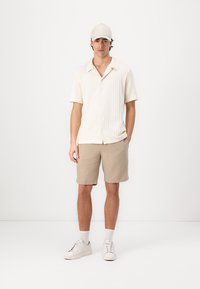 Krämfärgad kortärmad, texturerad skjorta med krage, tillsammans med ljusbeiga shorts. Outfitten inkluderar vita sneakers och en ljus keps.