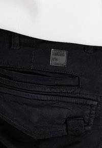 Nahaufnahme der Gesäßtasche einer schwarzen Hose mit Alpha Industries Logo-Patch und Gürtelschlaufe aus schwarzem Stoff.