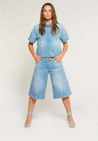 Chemise en denim à manches courtes, devant à boutons, et deux poches poitrine, associée à des culottes en denim arrivant au genou en bleu clair. Talons avec lanières inclus.