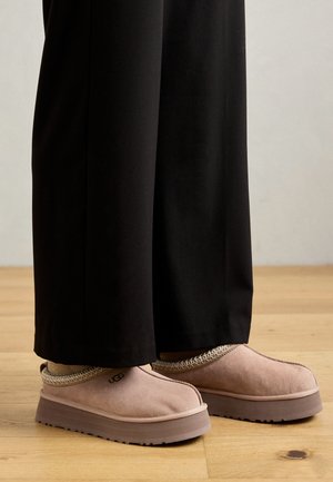 Des chaussures slip-on en suède rose avec un accent tissé texturé et une épaisse semelle marron, assorties à un pantalon noir sur un sol en bois.