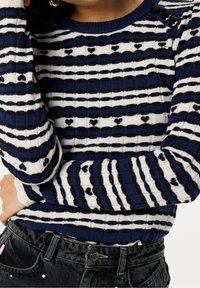 Námořnický a bílý pruhovaný pletený svetr se světlými vzory srdcí, ribbovanou texturou a černými mašlemi na ramenou. Vytvořeno v kombinaci s tmavými džíny.