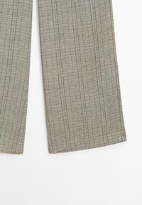 Gros plan d’un pantalon beige avec un motif à grille noire subtile et des jambes larges et droites sur un fond blanc.