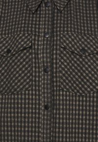 Flanellskjorta i mörkgrått och beige rutmönster, med spetsig krage, två ficklock och svarta knappar längs framsidan.