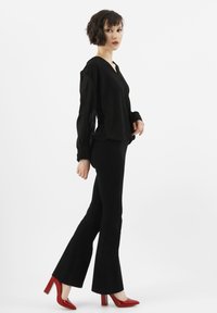 Zwarte transparante blouse met lange mouwen, gecombineerd met uitlopende zwarte broek en rode puntige hakken. De silhouet is op maat gemaakt en modern.